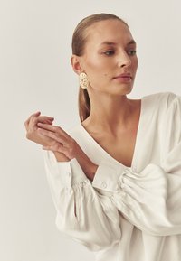 Bela bluza z dolgimi rokavi in volumnimi nabranimi rokavi. Veliki zlati ovalni uhani z teksturo poudarjajo eleganten videz. Naraven, gladek ton kože.