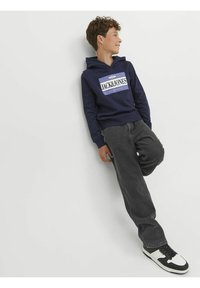 Sudadera azul marino con logo blanco, cordones y dobladillo acanalado. Combinada con jeans gris oscuro y zapatillas negras con detalles en blanco.
