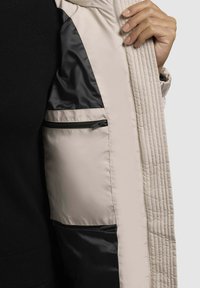 Hele halli polsterdatud jakk, millel on tikandiga disain ja must sisepokk, lukuga, et turvaliselt asju hoida.