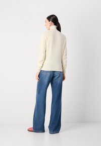 Un pull beige clair à col haut avec des manches longues, associé à un jean en denim bleu ample. La tenue présente des poignets côtelés et une coupe décontractée.