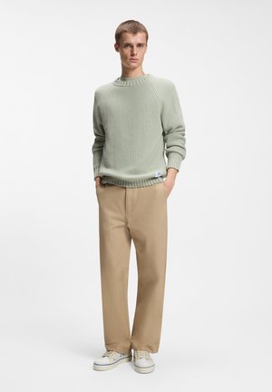 Νέος άνδρας με ανοιχτό πράσινο πλεκτό πουλόβερ, μπεζ παντελόνι και λευκά αθλητικά παπούτσια, όρθιος με τα χέρια στις τσέπες σε απλό λευκό φόντο.