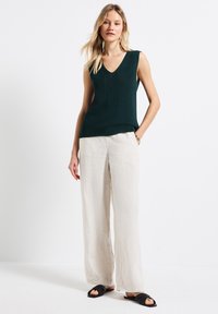 Bezrękawnik w serek w ciemnozielonym kolorze z dzianiny, w połączeniu z jasnobeżowymi spodniami typu wide-leg z lnu i czarnymi sandałami wsuwanymi.