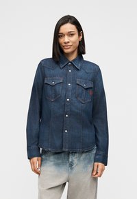 Γυναίκα με ίσια μαύρα μαλλιά, φορούσα σκούρο μπλε τζιν πουκάμισο με κουμπιά και ανοιχτόχρωμα τζιν παντελόνια, στέκεται μπροστά σε λευκό φόντο.