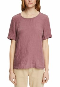 Femme portant un chemisier mauve uni à manches courtes et un pantalon beige, avec une expression neutre et une posture détendue.