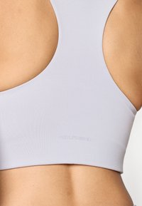 Sostén deportivo de color lavanda claro con un diseño de corte alto y espalda abierta. Logo grabado en la parte inferior. Tela suave y elástica con costuras mínimas.