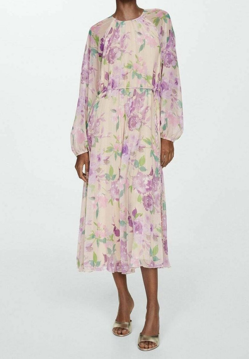 Femme portant une longue robe fleurie transparente avec des motifs violets et verts et des manches longues, associée à des talons ouverts dorés.