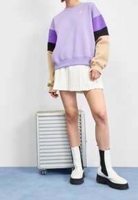 Sudadera morada con mangas negras, azules y beiges, combinada con una falda blanca con pliegues y botas de tobillo en blanco y negro sobre un suelo azul claro.