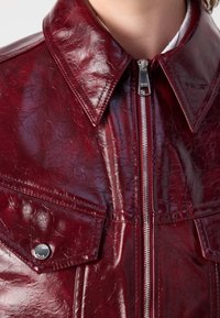 Czerwona kurtka z sztucznej skóry o błyszczącym wykończeniu, z zakończonymi w szpic kołnierzykiem, przednim zamkiem błyskawicznym i dwoma kieszeniami na klatce piersiowej, o pofalowanej fakturze.