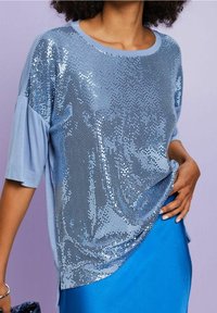 Mujer vistiendo una blusa corta de lentejuelas azul claro y una falda azul brillante, posando con una mano en la cadera contra un fondo púrpura.