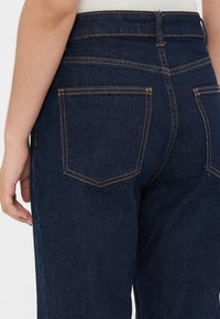 Vista aproximada de uma pessoa a usar jeans de ganga azul escuro, mostrando os bolsos traseiros e parte de uma blusa branca.