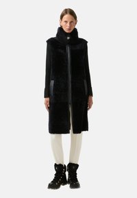 Kamizelka z sztucznego futra w ciemnym granacie, o długości do kolan, z wysokim kołnierzem, zamkiem błyskawicznym i bocznymi kieszeniami. Połącz ją z beżowymi legginsami i czarnymi butami ocieplanymi futrem.