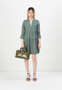 Robe verte à volants avec un décolleté en V et des manches trois-quarts, associée à un sac à main vert foncé avec un détail en étoile et des sandales à pois.