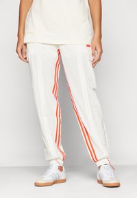 Alavartalo pukeutuneena valkoisiin urheiluhousuihin, joissa on oranssit raidat, ja valkoisiin lenkkareihin, joissa on kumipohjat, seisoo yksinkertaista valkoista taustaa vasten.