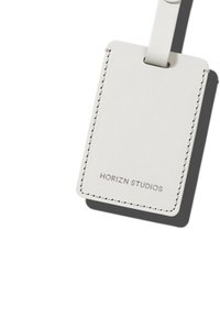 Weißes Ledergepäcketikett mit abgerundeten Kanten, sichtbaren Nähten und einem geprägten Logo "HORIZN STUDIOS" unten. Enthält eine Schlaufe zur Befestigung.