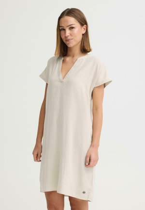 Jonge vrouw met stijlbruin haar, gekleed in een korte mouw, beige, knielange jurk met een V-hals, staand tegen een effen achtergrond.