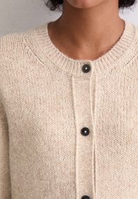 Beige gebreide cardigan met een ronde hals en zwarte knopen. De stof lijkt zacht met een gezellige, gestructureerde afwerking.