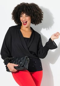 Schwarze Wickelbluse mit Puffärmeln, kombiniert mit einem enganliegenden schwarzen Top mit kleinen Nieten. Helle rote Hosen und eine gesteppten schwarzen Handtasche.