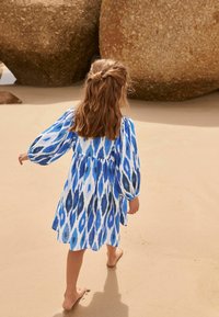 Modrá a bílá šaty s vzorováním, s nabíranými rukávy, volný střih a délka ke kolenům. Stylizované pro plážové prostředí, písek a velké kameny v pozadí.