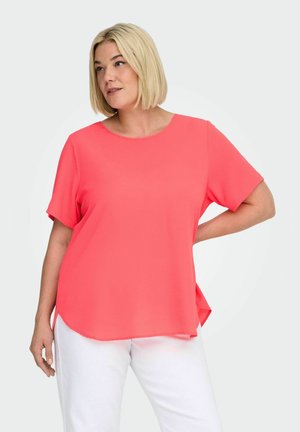 Kobieta o blond krótkiej fryzurze bob, ubrana w koralową bluzkę z krótkim rękawem i białe spodnie, pozująca z jedną ręką na biodrze na jednolitym tle.