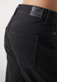 Jeans de mezclilla negra con un acabado texturizado, que presentan un bolsillo trasero cosido visible y una etiqueta de logo negra en la cinturilla.