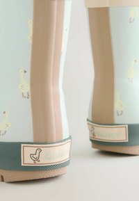 Hellblaue Gummistiefel mit beigen Akzenten, versehen mit einem Entenmuster und einer strukturierten grünen Sohle. "Quack"-Etikett an der Ferse.