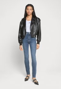 Veste de biker en cuir synthétique noire avec larges revers, fermeture éclair et poignets côtelés, associée à un jean en denim bleu ajusté et des chaussures noires à bout pointu.