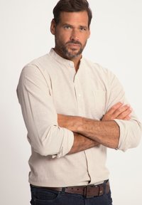 Helles beiges Button-up-Hemd mit langen Ärmeln, ausgestattet mit einem Stehkragen und einer Tasche. Getragen mit dunklen Jeans und einem braunen Gürtel.