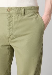 Light olive green chinos with a smooth texture, featuring a button closure, side pockets, and straight legs. Focus on functional design details.