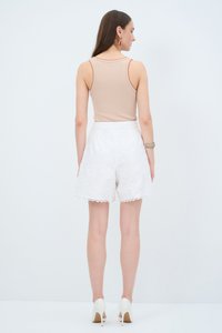 Top sin mangas beige con ribete naranja, combinado con pantalones cortos blancos bordados que presentan un dobladillo en forma de concha, y zapatos de tacón blancos.