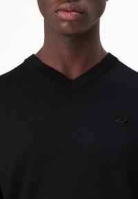 Maglione nero a collo a V in texture a coste, con un piccolo accento del logo scuro sul lato sinistro. Il tessuto appare morbido e aderente.