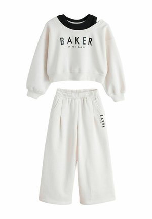 Person iført cremefarvede joggingbukser med hånd i lommen og sort lodret "BAKER BY TED BAKER" tekst på buksebenet.