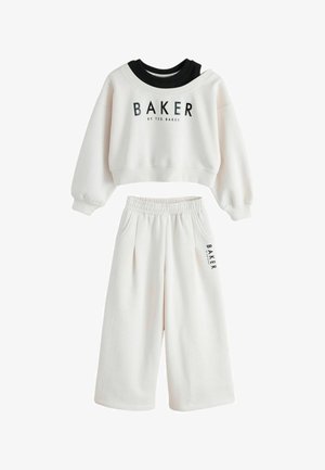 Person iført cremefarvede joggingbukser med hånd i lommen og sort lodret "BAKER BY TED BAKER" tekst på buksebenet.