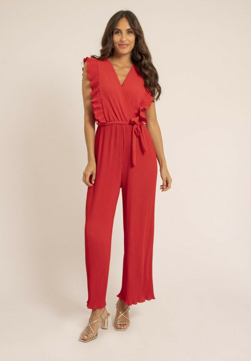 Combinaison rouge plissée avec des manches à volants, un décolleté enroulé et une taille nouée. Accentuée par des jambes de pantalon évasées et portée avec des talons en argent à lanières.