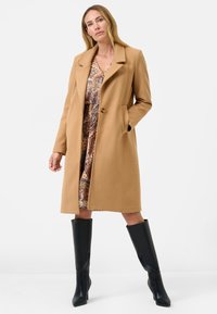 Camelkleurige jas met een gestructureerd ontwerp, twee zijzakken en een enkele knoopsluiting, gelaagd over een patroonjurk en gecombineerd met zwarte kniehoge laarzen.
