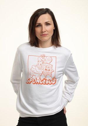 Donna con capelli castani fino alle spalle che indossa una felpa bianca con grafica arancione di SpongeBob e Patrick e pantaloni neri.