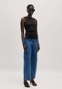 Schwarzes ärmelloses Oberteil mit seitlicher Raffung, kombiniert mit blauen weit geschnittenen Jeans und schwarzen High Heels, vor einem neutralen Hintergrund.