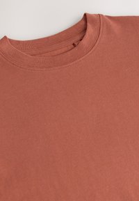 Maglietta in cotone color ruggine con collo a girocostina, texture liscia e dettagli di cuciture precise. Design minimalista senza grafiche visibili.