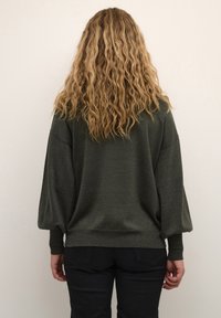 Maglione verde scuro con vestibilità morbida, maniche ampie e polsini a coste. Materiale lavorato a maglia testurizzato, orlo posteriore più lungo. Pantaloni neri visibili.