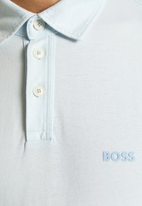 Polo shirt bleu clair à la texture douce, trois boutons et un col brodé. Présente un logo "BOSS" bleu clair sur la poitrine.