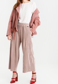 A woman stands wearing a pink suede jacket, white top, and pleated beige trousers, complemented by striking red block-heeled shoes.