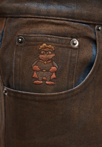 Καφέ υφασμάτινο denim με υφασμένη υφή, με κεντημένο καρτούν χαρακτήρα στην πίσω τσέπη, περιβαλλόμενο από ασημένια διακοσμητικά στοιχεία.