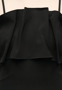 Μαύρο αμάνικο φόρεμα με φαρδιά, στρωματοποιημένη βολάν λεπτομέρεια στον θώρακα και λεπτούς ιμάντες στους ώμους σε λευκό φόντο.