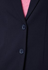 Marinblå blazer med två svarta knappar, slät tyg, skräddarsydd design och en subtil lyster, lager på en rosa texturerad tröja.