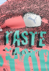 Gros plan sur un tissu rose et bleu sarcelle avec des points noirs texturés, des lettres sarcelles lisant partiellement "TASTE", et un logo Puma argenté.