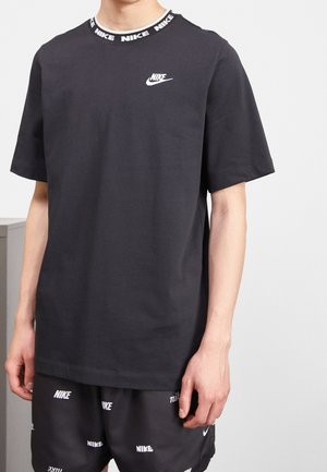 Mand iført sort Nike-t-shirt med hvidt logo på brystet og gentaget Nike-tekst på kraven, kombineret med sorte Nike-shorts med hvide logoer.