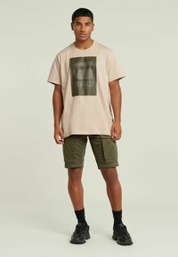 T-shirt beige chiaro con un design grafico verde scuro, abbinato a pantaloni cargo verde oliva. Il modello indossa calze nere e sneakers.