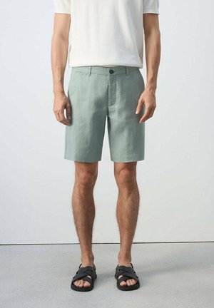 Hombre con pantalones cortos verde claro hasta la rodilla, camisa blanca de manga corta y sandalias negras, de pie frente a un fondo claro y liso.