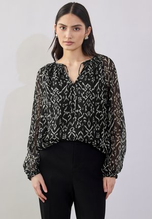 Blouse à motifs noir et blanc en tissu léger et transparent, avec un décolleté en V et des poignets froncés, portée sur un pantalon noir.