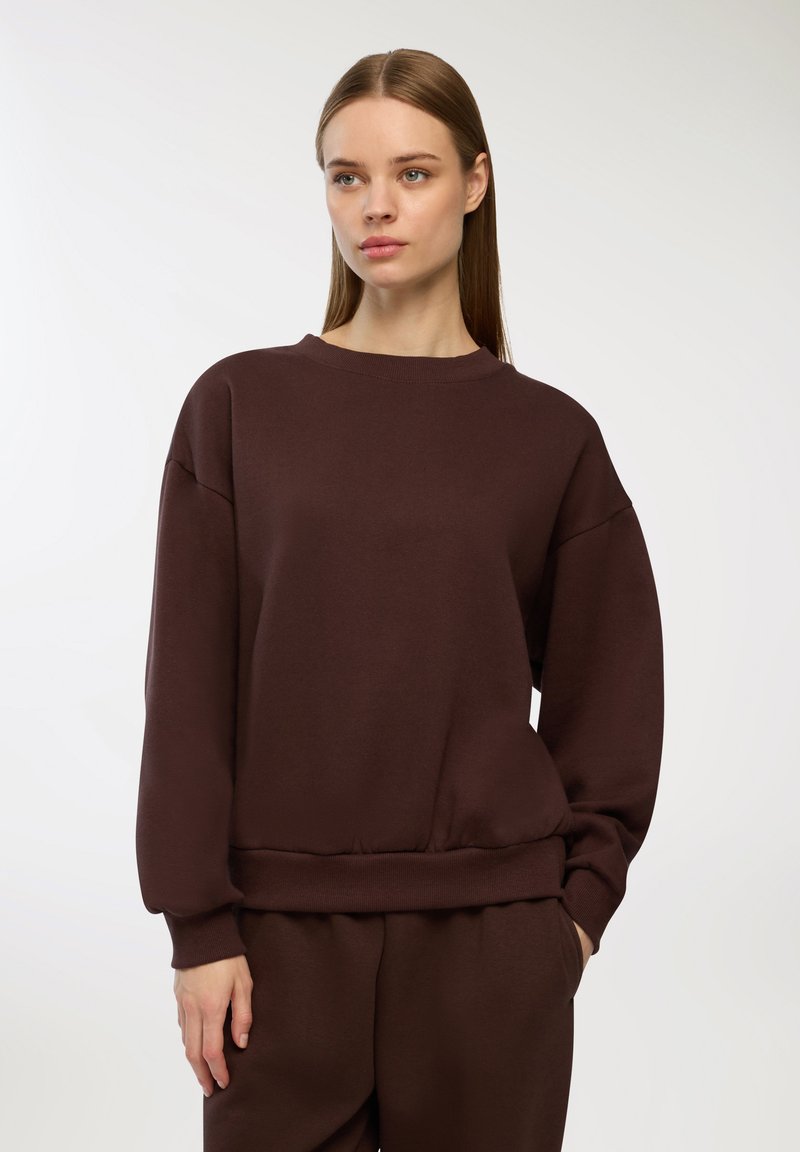 Brązowy, lekki sweter z okrągłym dekoltem, opadającymi ramionami i prążkowanymi mankietami. Posiada luźny krój i subtelną fakturę materiału.