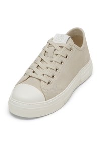 Sneaker in tela beige con punta in gomma bianca, lacci piatti, occhielli in metallo e un'etichetta di marca sulla linguetta. Presenta una superficie texturizzata.